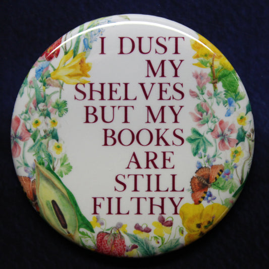 Button badge with floral design and text 'I dust my shelves but my books are still filthy' on a dark background