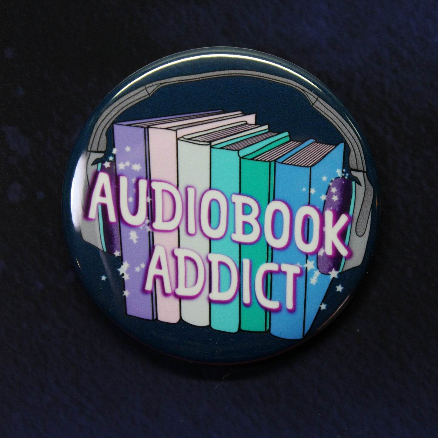 A round pinback button badge featuring a colorful illustration of books with headphones and the text 'AUDIOBOOK ADDICT' in pink, displayed on a dark textured background.