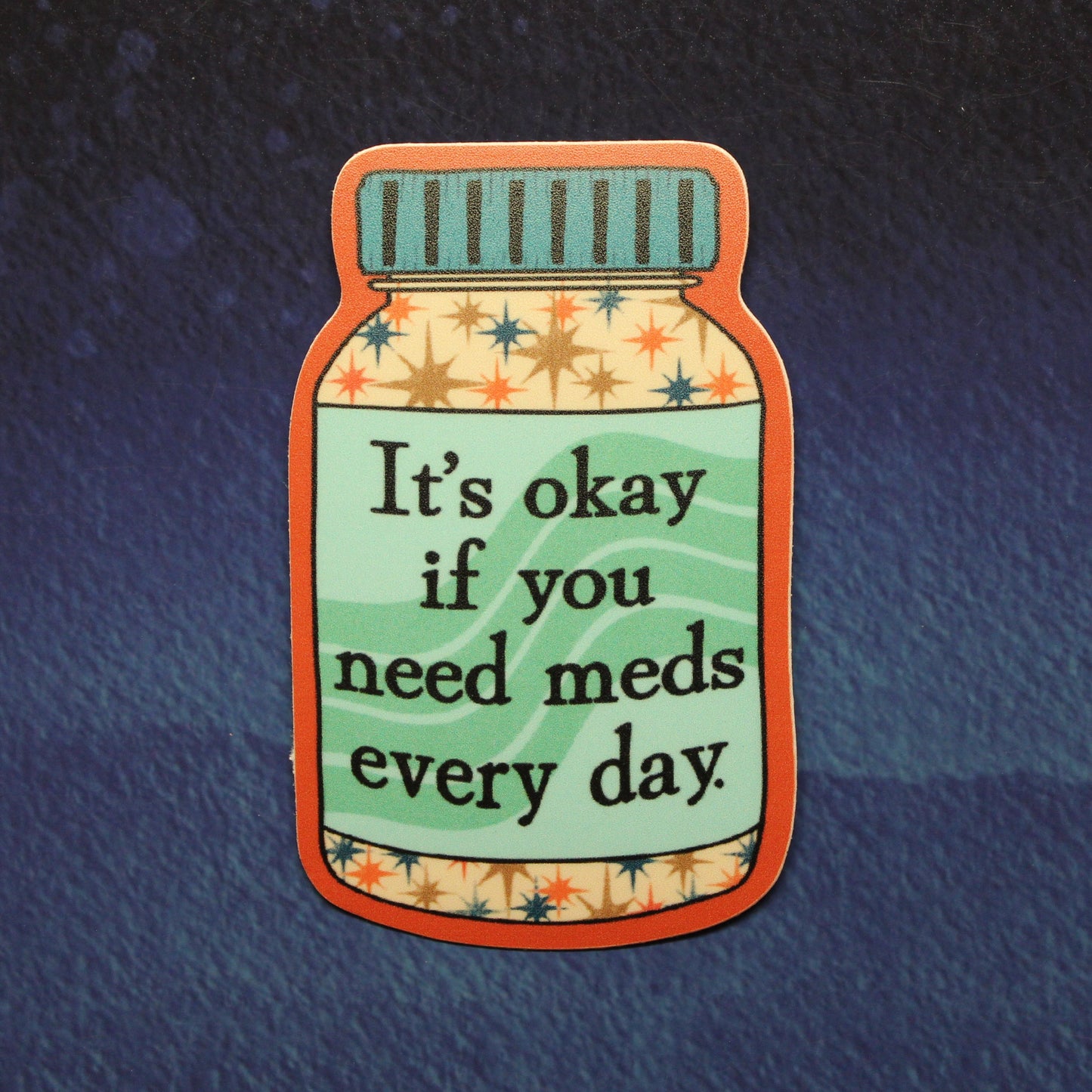 A die-cut sticker shaped like a pill bottle with the text 'It's okay if you need meds every day' in black lettering, set against a dark textured blue background.