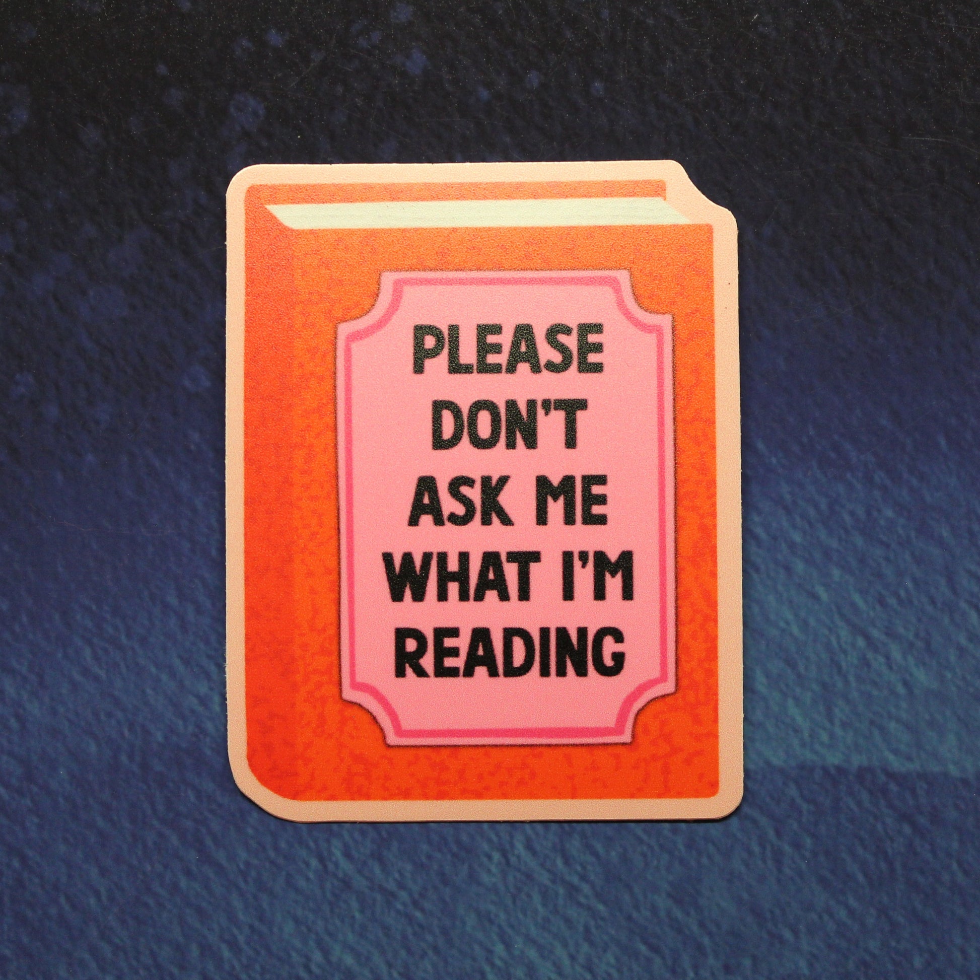 A rectangular book-shaped sticker or pin with a peach-orange cover and a pink title panel reading 'PLEASE DON'T ASK ME WHAT I'M READING' in black text, shown on a dark blue textured background.