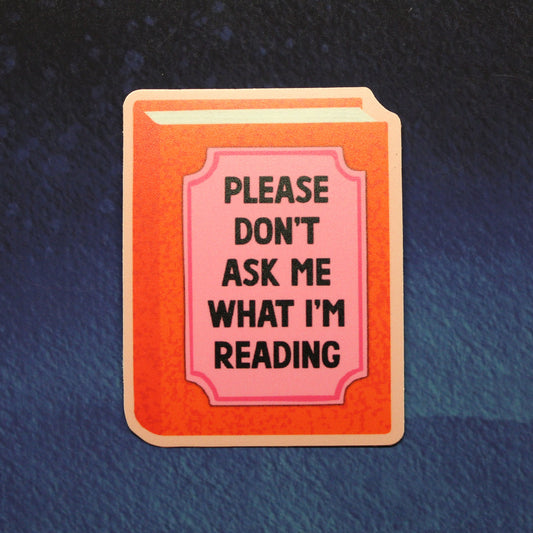A rectangular book-shaped sticker or pin with a peach-orange cover and a pink title panel reading 'PLEASE DON'T ASK ME WHAT I'M READING' in black text, shown on a dark blue textured background.