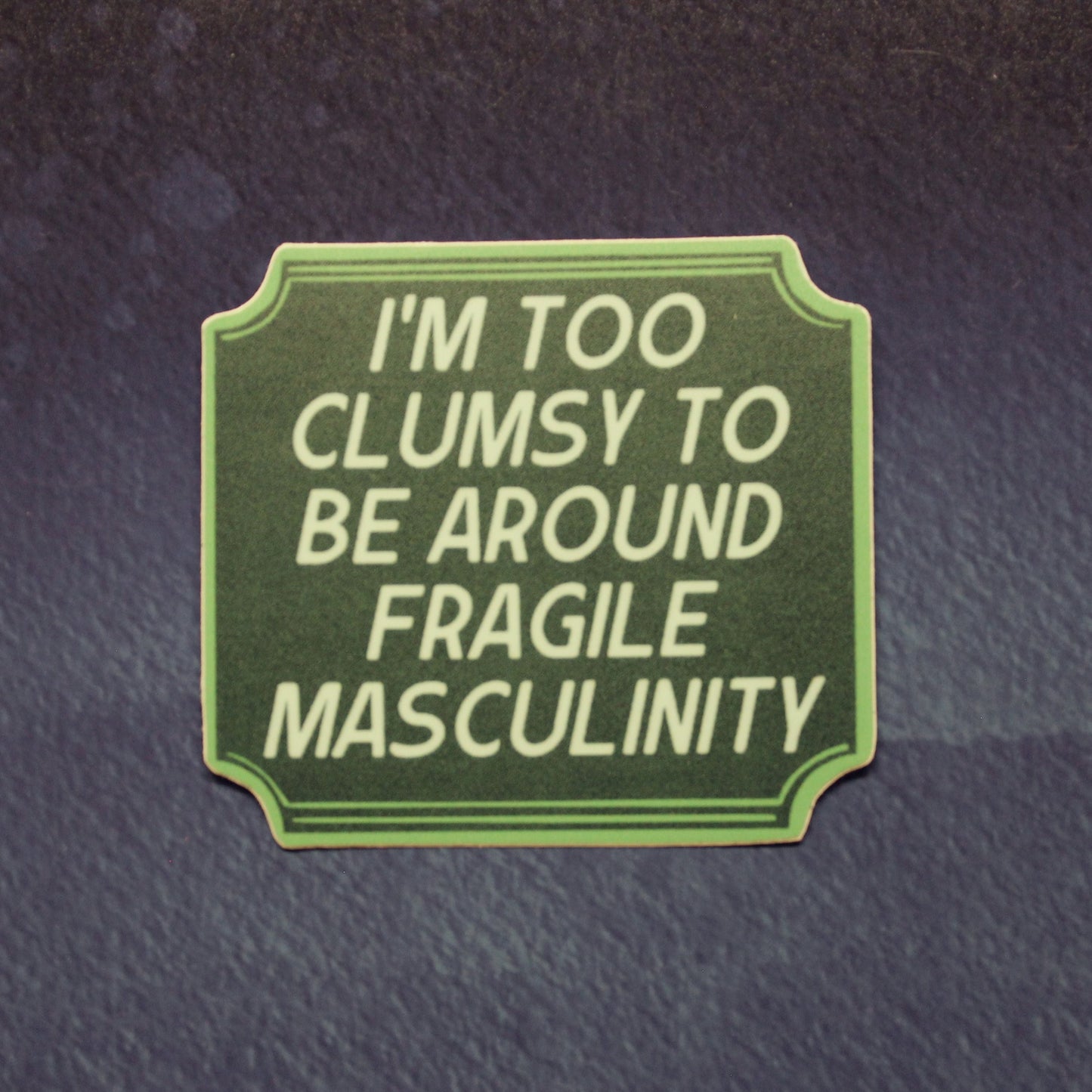 A rectangular green patch with rounded corners and decorative border, featuring the text 'I'M TOO CLUMSY TO BE AROUND FRAGILE MASCULINITY' in light green lettering, shown on a dark textured background.