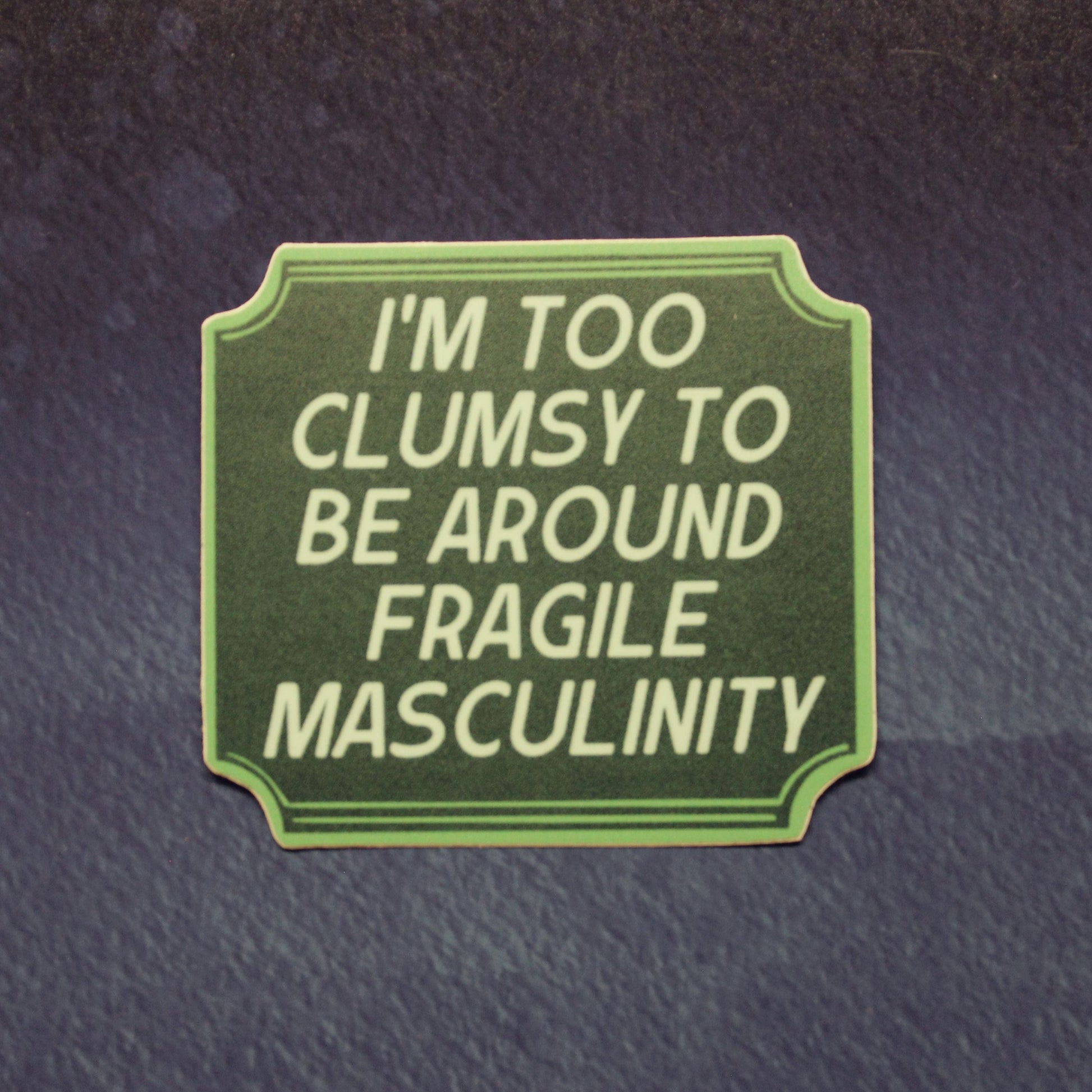 A rectangular green patch with rounded corners and decorative border, featuring the text 'I'M TOO CLUMSY TO BE AROUND FRAGILE MASCULINITY' in light green lettering, shown on a dark textured background.