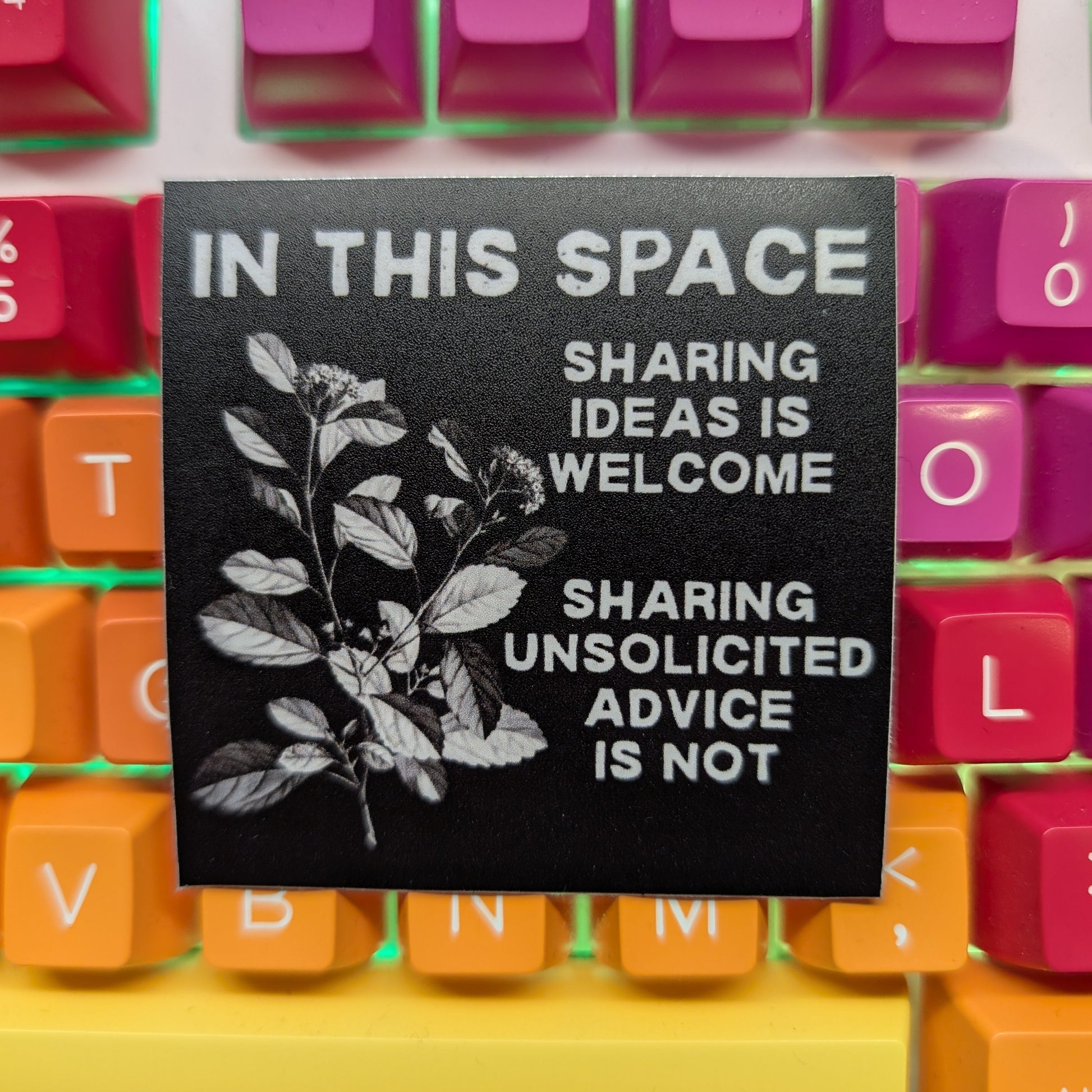 A black square desk mat with white text and botanical leaf graphics, placed on top of a colorful mechanical keyboard. The desk mat reads: 'IN THIS SPACE SHARING IDEAS IS WELCOME SHARING UNSOLICITED ADVICE IS NOT'.