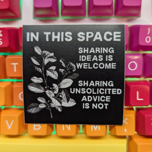 A black square desk mat with white text and botanical leaf graphics, placed on top of a colorful mechanical keyboard. The desk mat reads: 'IN THIS SPACE SHARING IDEAS IS WELCOME SHARING UNSOLICITED ADVICE IS NOT'.