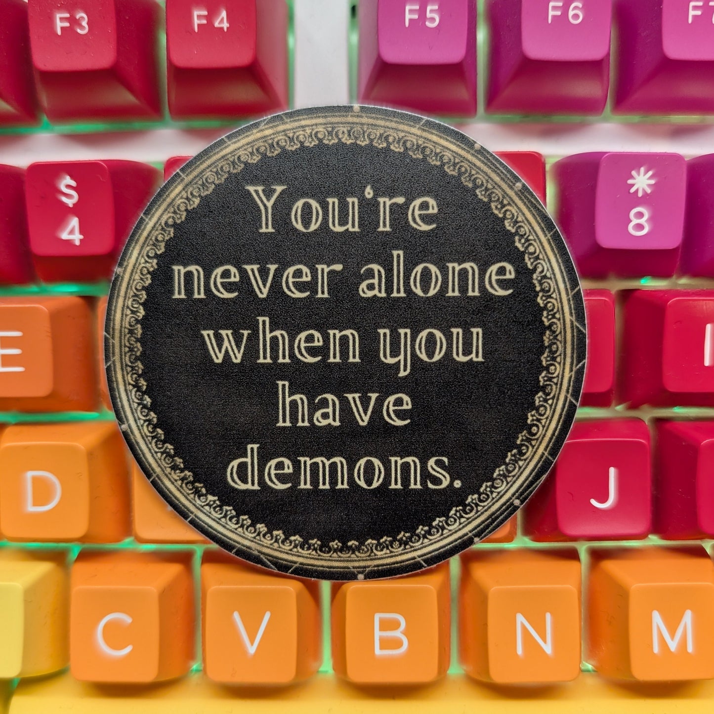 A round black coaster with gold lettering that reads 'You're never alone when you have demons.' placed on top of a colorful mechanical keyboard with pink, orange, and yellow keys.